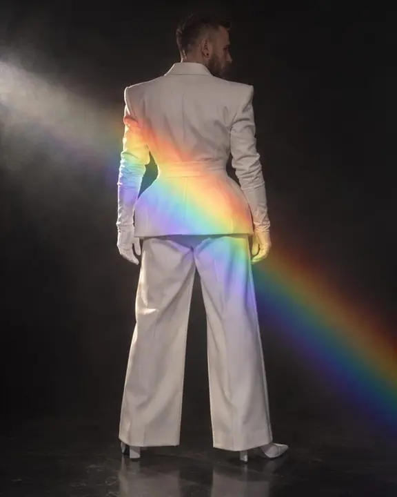 Back view of a man in a white padded-hip blazer, a spectrum band falling across the hips in a dark room