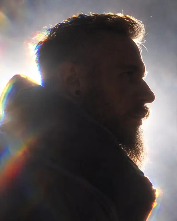 Profile of a bearded man in darkness, forehead, nose and beard traced by a thin rainbow rim of backlight