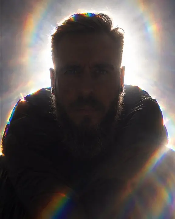 Frontal view of a man with light behind his head forming a rainbow halo, his eyes barely visible in shadow