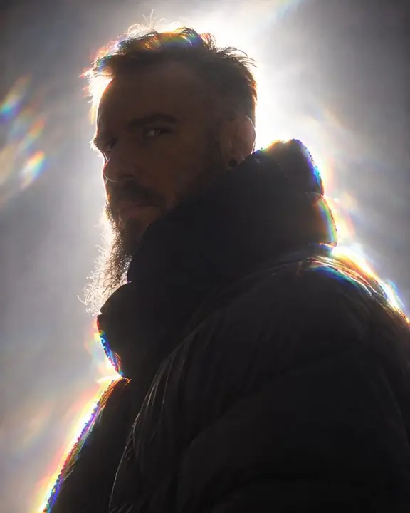 Three-quarter view of a bearded man in a funnel collar split into colors at the edge, one eye catching light