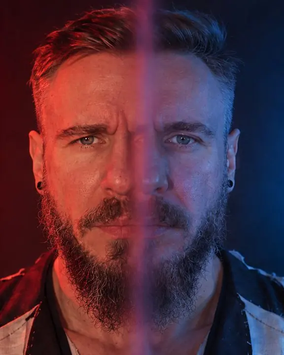 Head and shoulders of a bearded man, his face split down the middle, one half red, the other a shifted blue copy