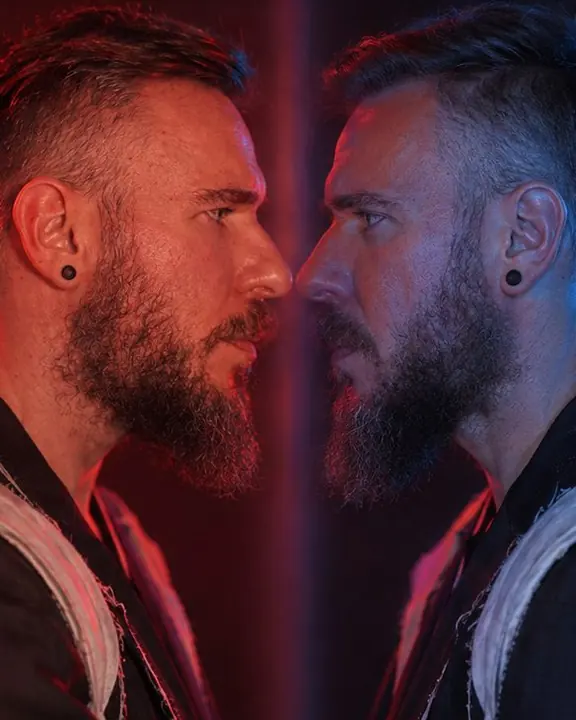 Close-up of two profiles of a bearded man nose to nose across a soft vertical seam, one red, one blue