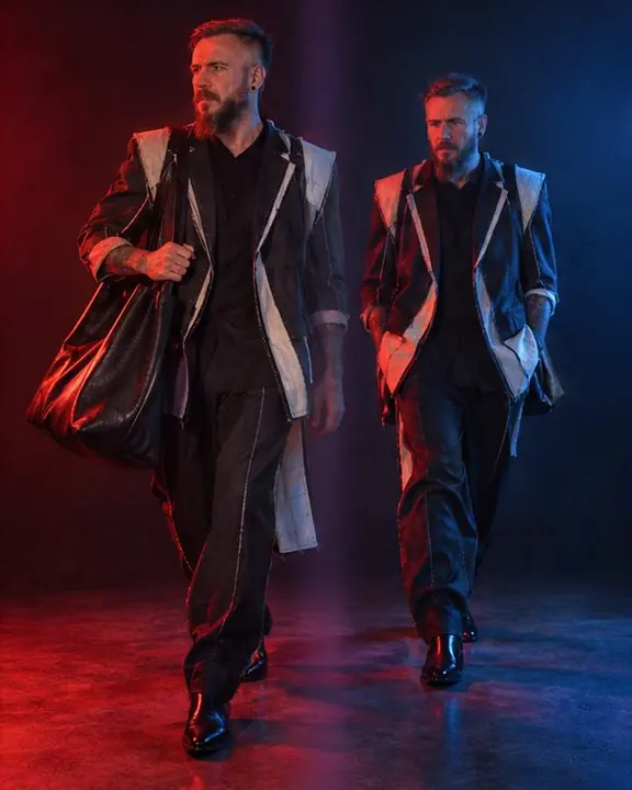 A man in an inside-out suit walking, his blue twin a step ahead of the red one against a dark background