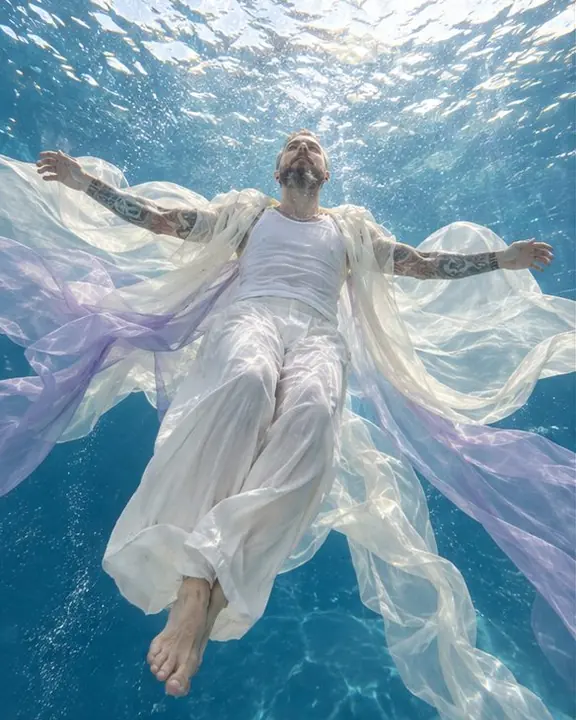 A bearded man floats horizontally beneath a bright pool surface, ivory fabrics spreading from him like wings