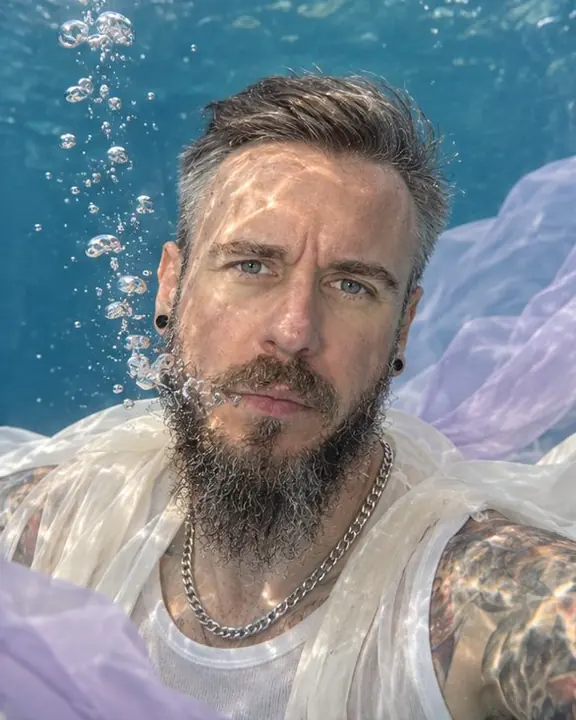 Waist-up underwater view of a calm bearded man, a thin stream of bubbles rising from his lips, caustics on his cheek