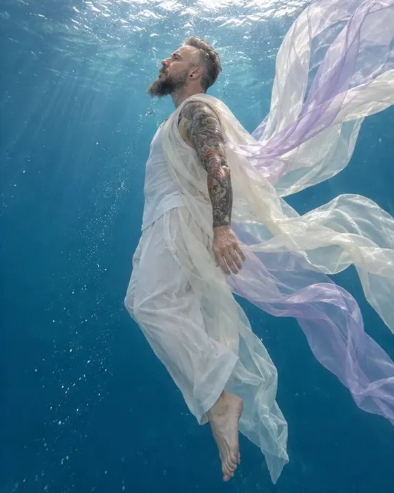Side view of a man drifting upright underwater, a sheer ivory coat billowing upward, toes pointed