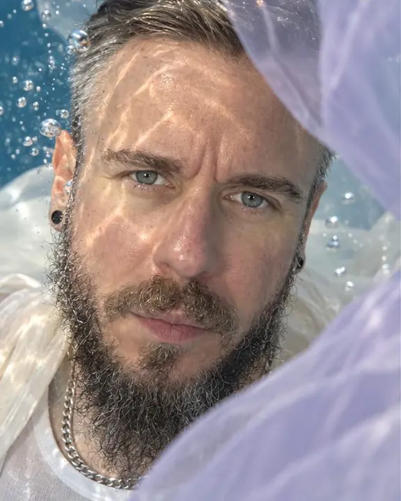 Close underwater portrait of a bearded man with eyes open to the lens, lilac chiffon drifting across the frame