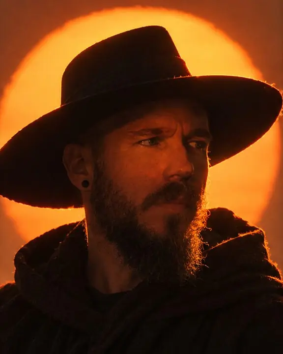 Head and shoulders of a man in a wide-brimmed hat against the sun, orange light touching his eyes