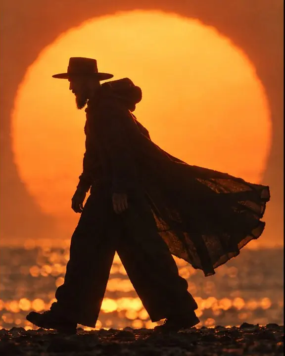A black silhouette of a man walking across a huge setting sun, his cape trailing behind in the haze