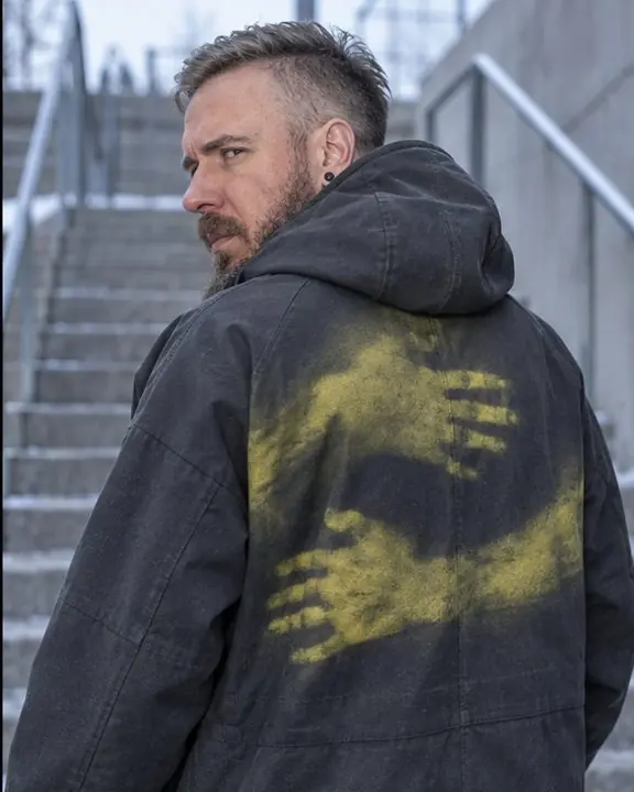 Back view of a man in a graphite hooded coat with a yellow print of an embrace, looking over his shoulder
