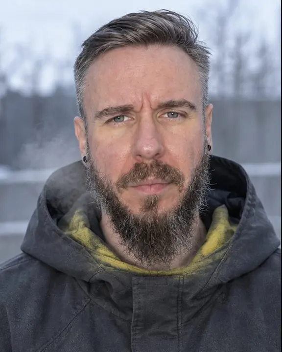 Head and shoulders of a bearded man, hood down, a warm yellow halo around the neckline, breath in cold air