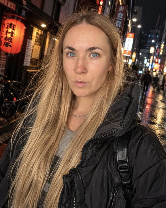 A woman with long blond hair, windblown, faces a bare flash in a black nylon jacket, red lanterns and a wet street behind