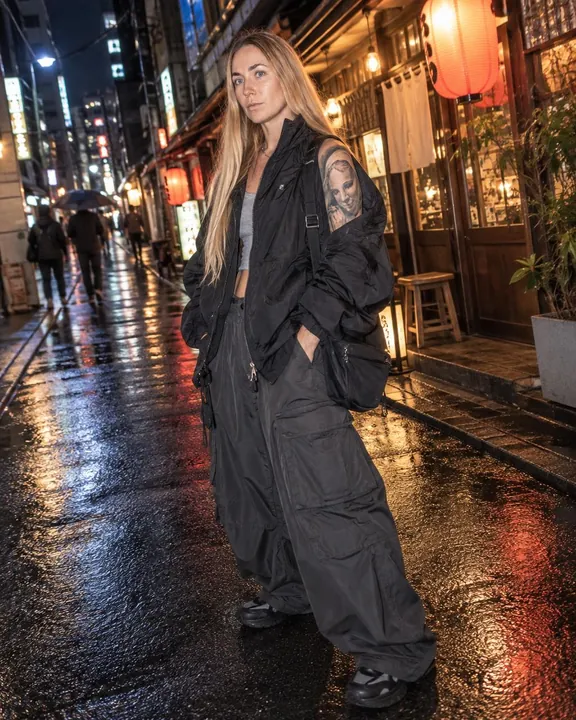 Full-length woman with long blond hair on wet asphalt, black nylon jacket off one shoulder, wide cargo trousers, shoulder bag