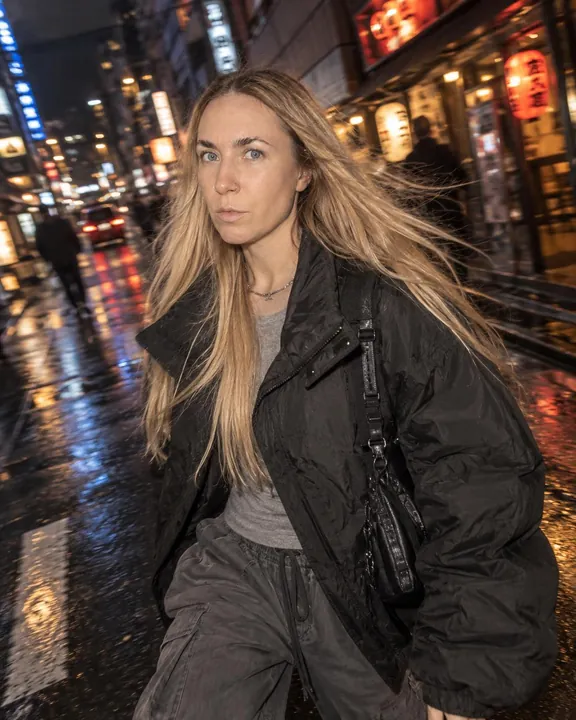 A woman with long blond hair strides across a wet night street, hair blowing, a black bag strap over her nylon jacket