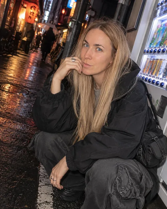 A woman with long blond hair crouches by a vending machine, chin on a hand with silver rings, a slight smile at the lens