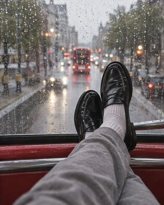 A man with his loafers up on the front rail of a bus top deck, rain on the glass and a blurred street ahead