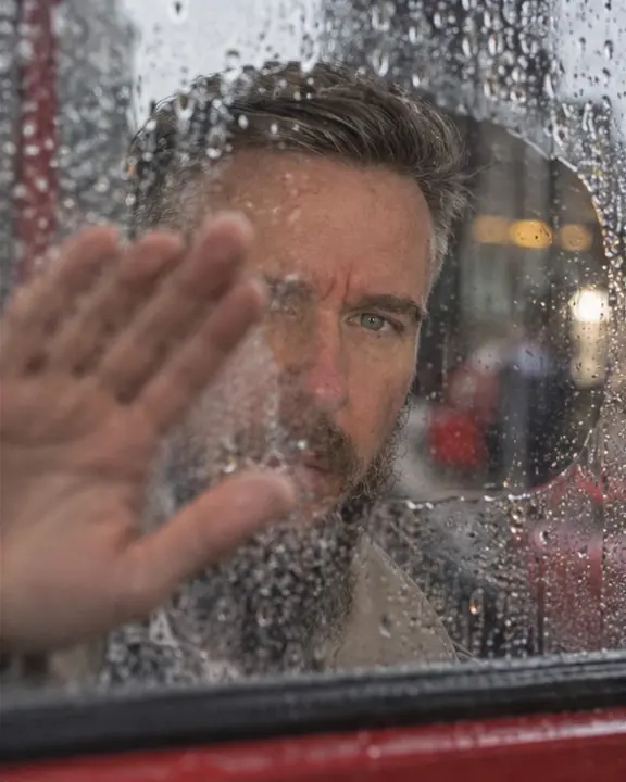 A hand wipes a clear patch on a steamed-up bus window, revealing a bearded man's eyes behind the glass