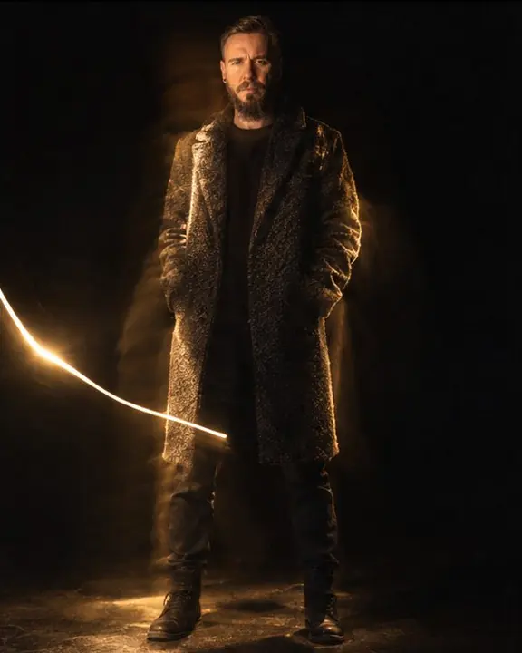 A man in a bouclé coat lit in uneven warm strokes, one bright flashlight streak crossing the dark frame