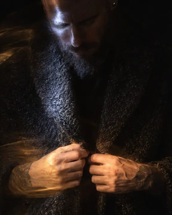 A man's hands and textured coat lit by flashlight, his face barely touched by a cold white stroke