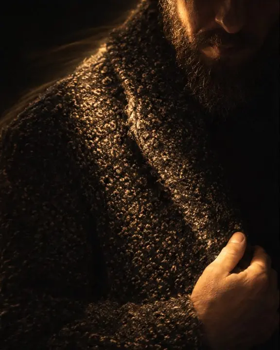 Textured bouclé coat fabric raked by a narrow stroke of warm flashlight light in total darkness
