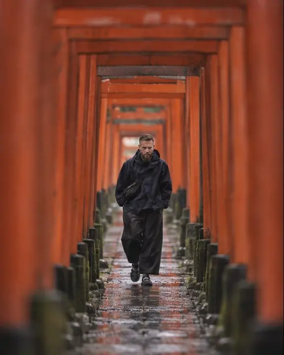 A bearded man stands deep inside a tunnel of vermilion torii gates, a dozen soft out-of-focus gates framing him