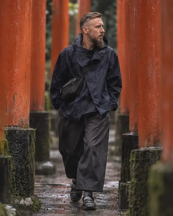 A man in an indigo nylon wrap jacket steps between two red torii gates, soft side light on the fabric
