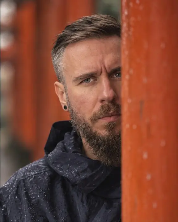 Close-up of a bearded man framed by one sharp vermilion pillar, softer red gates blurred behind him