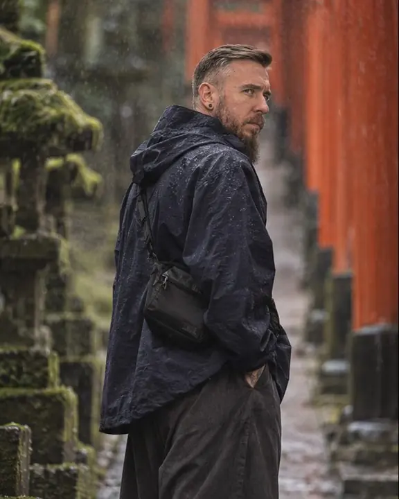 A bearded man looks back over his shoulder in light drizzle, dark moss and red gates behind him in Kyoto