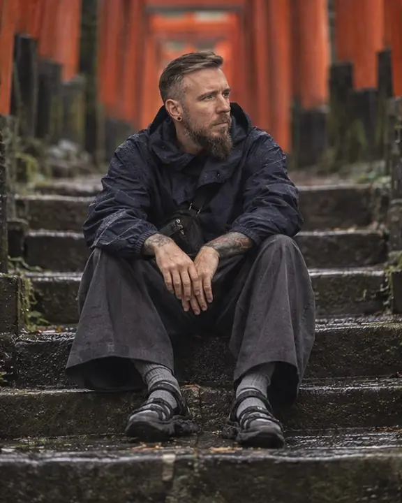 A man in indigo and charcoal sits on old stone steps with rows of vermilion torii gates rising behind