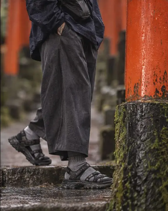 Indigo technical nylon against a wet vermilion torii pillar streaked with green moss in soft overcast light
