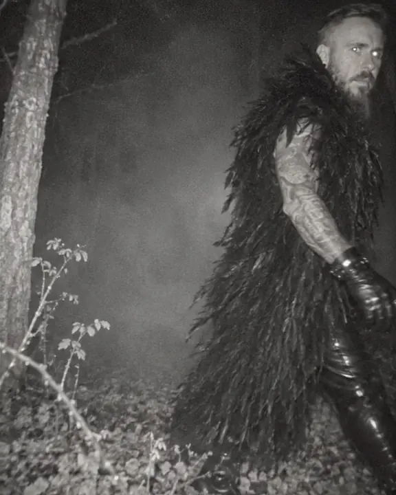 Grainy night-vision view of a bearded man striding past a camera trap, half out of frame, eyes catching the flash