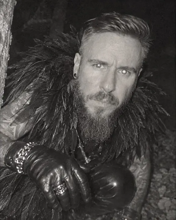 A bearded man crouches close to a forest trail camera, curious, his face filling half the infrared frame