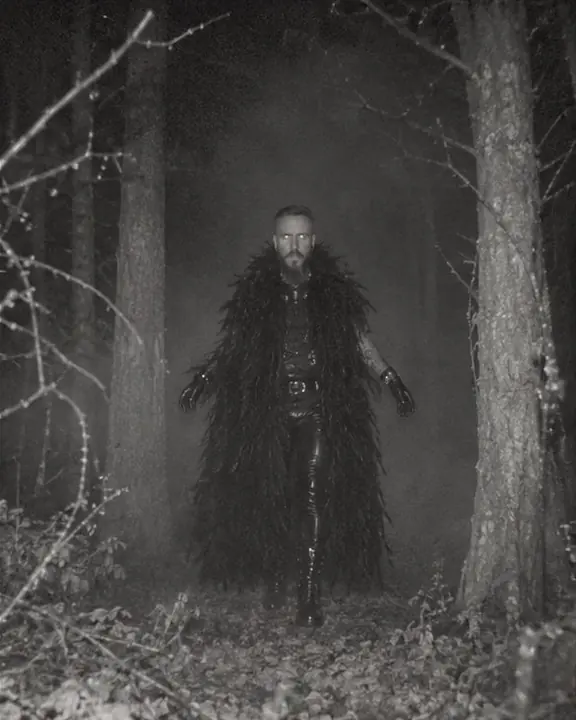 Distant full-length of a man in a black feather coat standing between foggy tree trunks at night