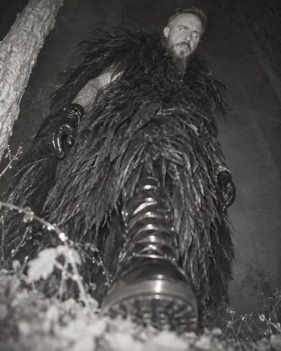 Low view of legs, patent boots and a feathered coat hem crossing the frame, a face looking down from above