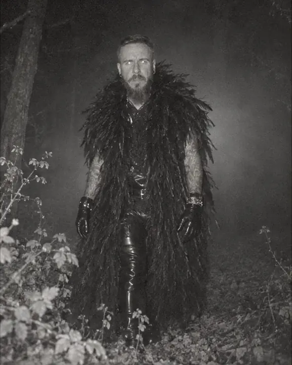 A bearded man looks straight at a trail camera from two meters, his eyes reflecting the infrared flash