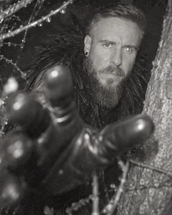 A black patent-gloved hand reaches toward a night camera, wet forest branches around it in gray-green