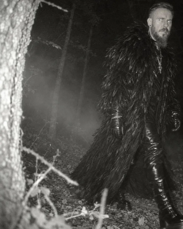 Grainy night-vision view of a bearded man striding past a camera trap, half out of frame, eyes catching the flash
