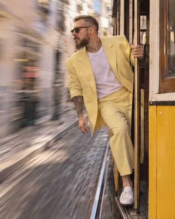A bearded man in a pastel-yellow suit holds the pole on a Lisbon tram step, the street streaking behind