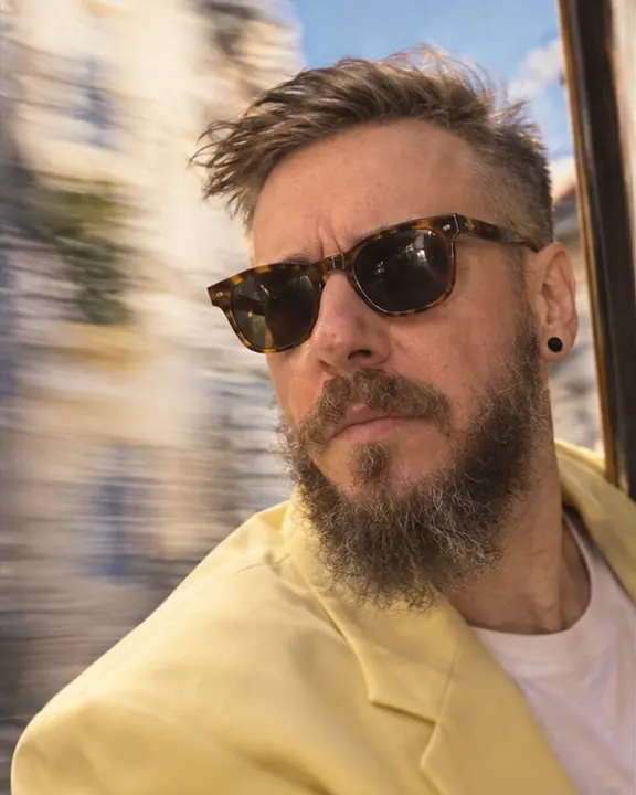 Close-up of a bearded man with wind in his hair, a warm pastel blur of Lisbon streets behind him