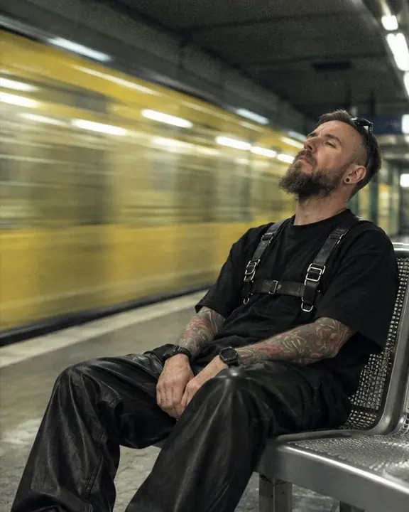A bearded man sits on a platform bench, head tipped back, a subway train streaking past in cold light