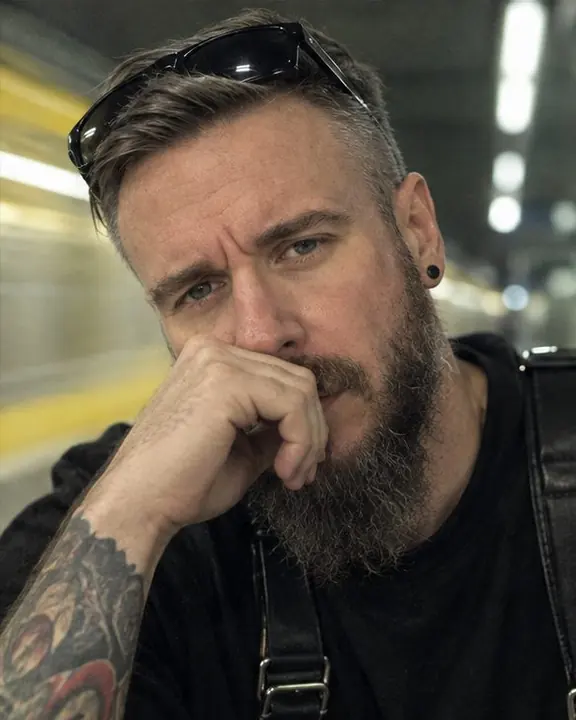 Close-up of a tired bearded man with shield sunglasses pushed up, lit by cold green-white fluorescent light