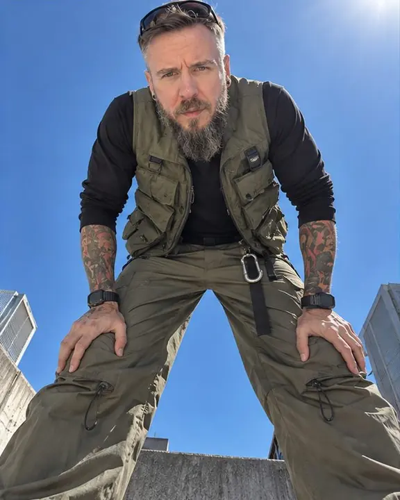 Straight-up view of a bearded man leaning over the camera with hands on knees, looking down, sky around his head