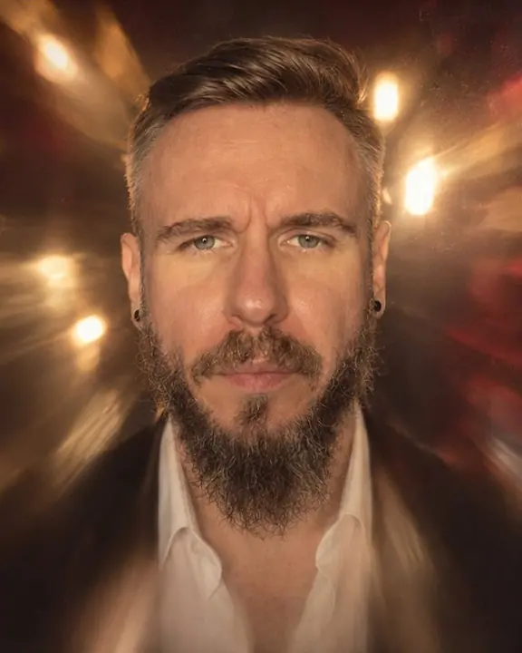 A bearded man's face in the clear center of the frame, bare bulbs behind him bursting into radial light streaks