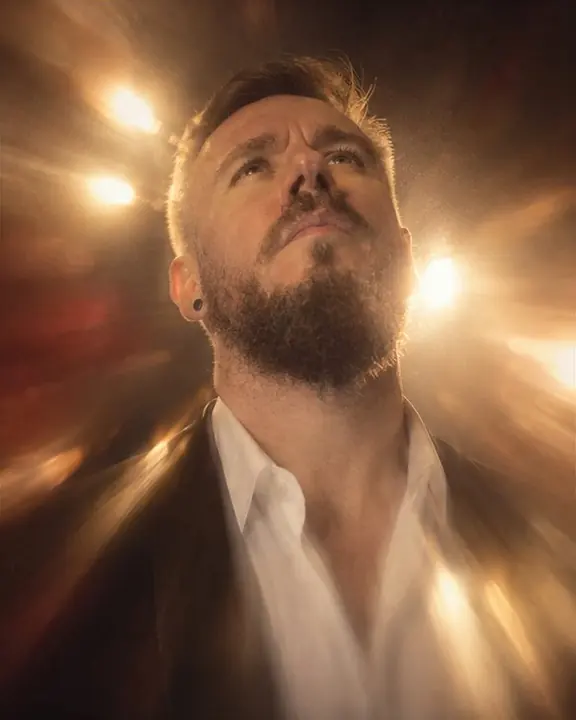 A bearded man tilts his head back, his white open shirt collar glowing and streaking in warm light