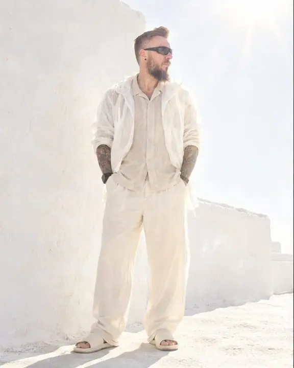 A bearded man stands still against a whitewashed wall in blazing sun, flare spilling from the top corner of the frame