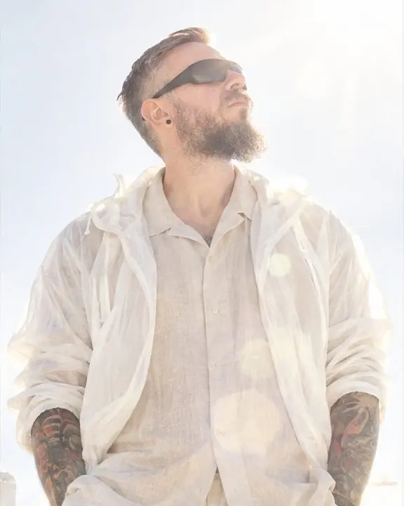 A man in black wraparound sunglasses tilts his head up into the sun, a translucent white rain shell glowing