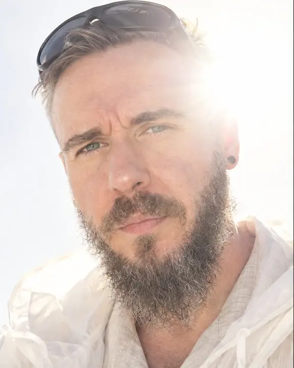Close portrait of a bearded man with sunglasses pushed up, squinting into the light, half his face dissolving in flare