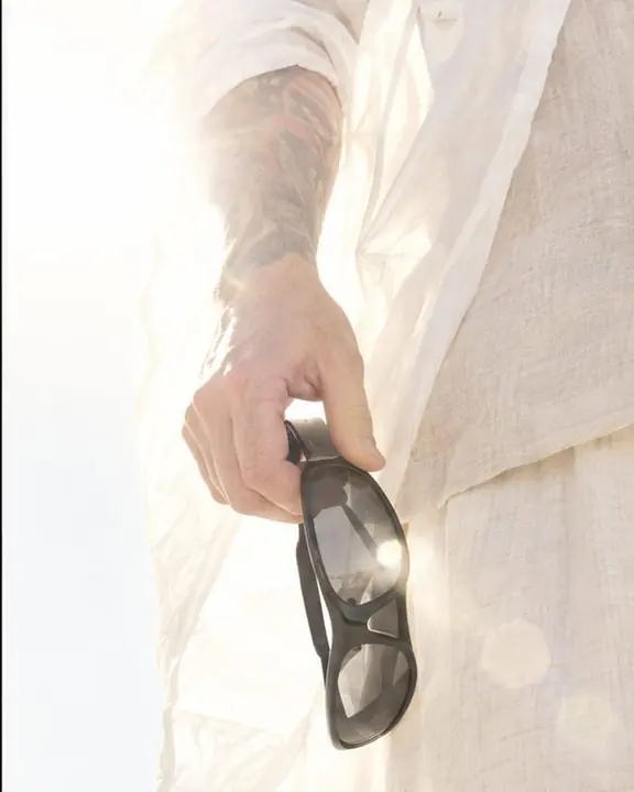 Close-up of a hand holding black sculptural sunglasses against a translucent white rain shell