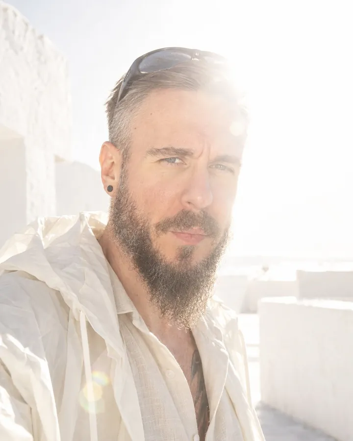 Close portrait of a bearded man with sunglasses pushed up, squinting into the light, half his face dissolving in flare