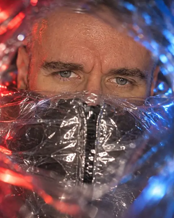 Close-up of a bearded man's sharp eyes above a plastic-wrapped collar glinting with crinkled highlights